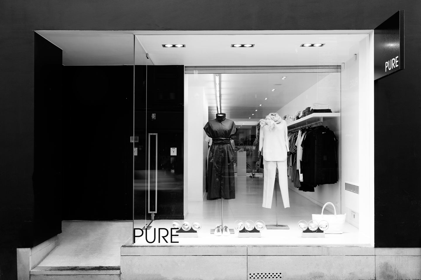 De etalage van Pure in de Voorstraat in Kortrijk, ’s avonds verlicht, met een jurk en een broekpak achter het glas